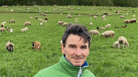 Uwe Becker steht vor einer Wiese, auf der zahlreiche Schafe und einige Ziegen grasen